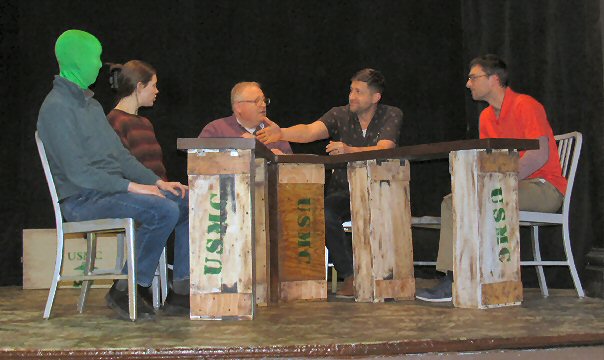 five people sitting around a makeshift deck.  the
head of one is covered by a green mask